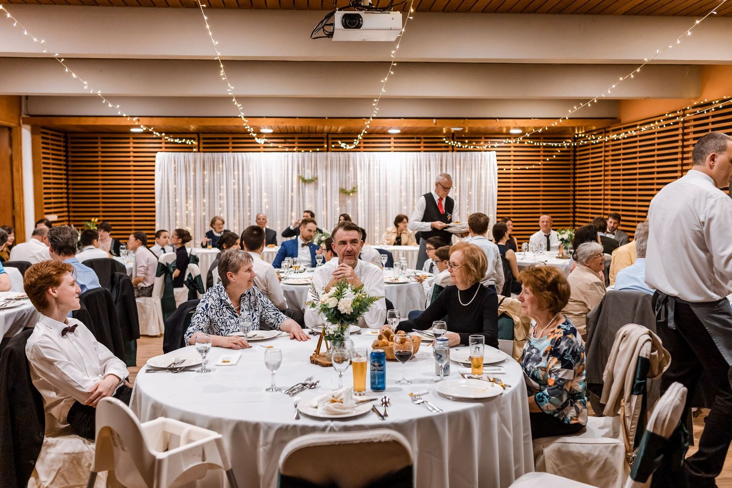 Panoramic view of a wedding reception venue with guests dining at round tables under warm lighting and decorative accents.