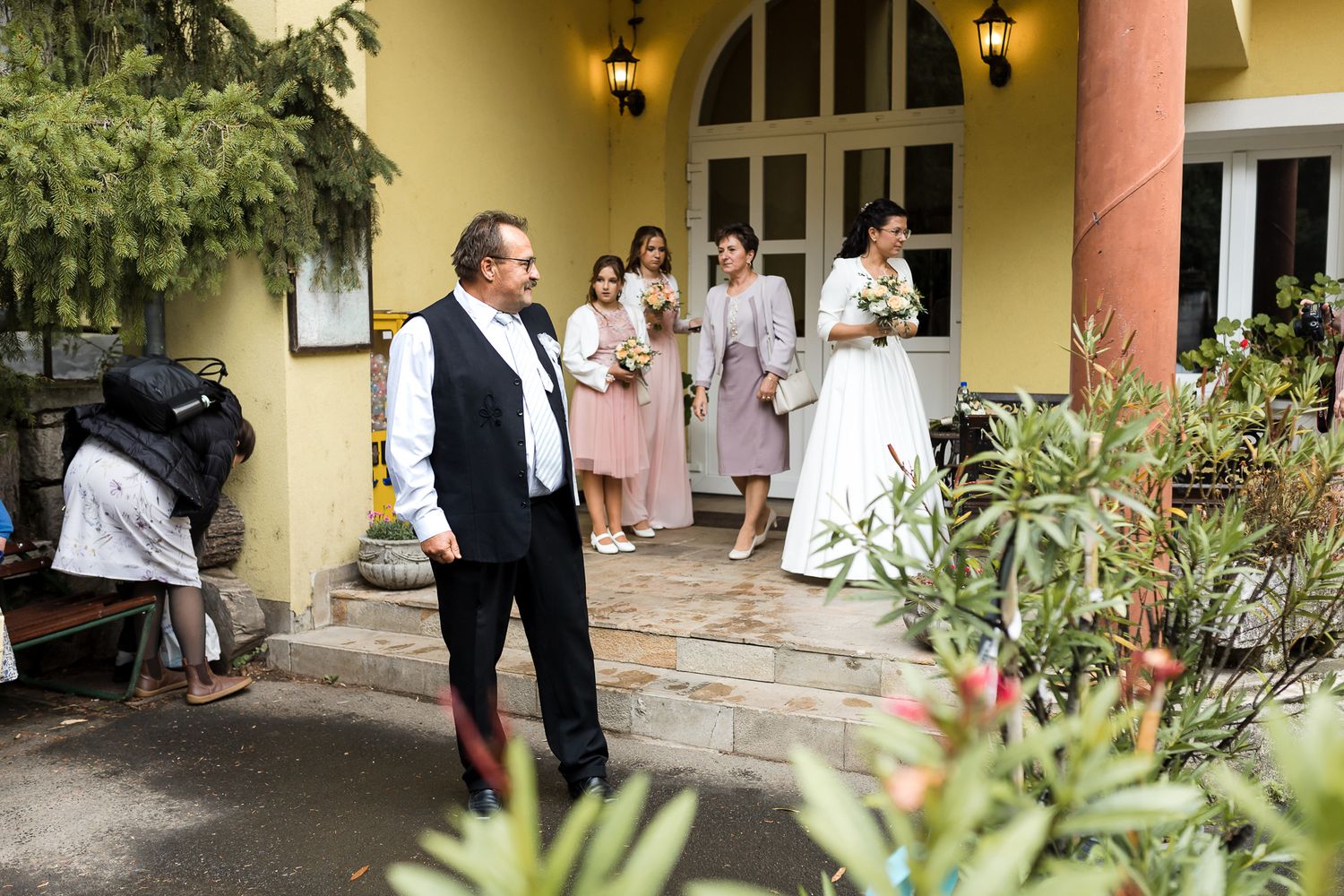Wedding guests gather outside a yellow Mediterranean-style venue with arched doorways and decorative wall sconces.