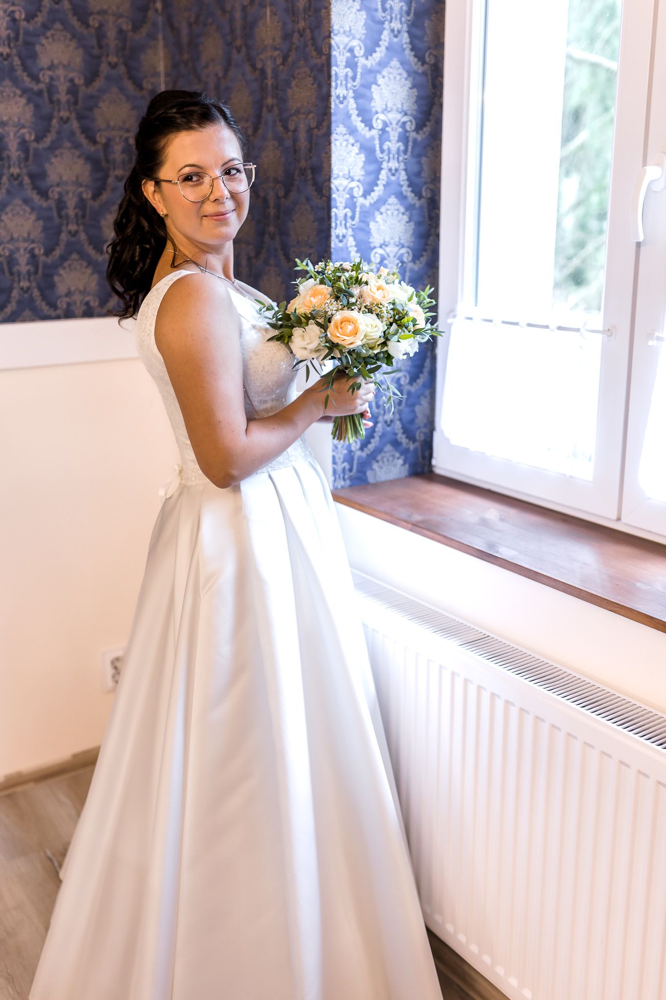 A bride in a classic white wedding dress stands by a window holding a delicate bouquet of flowers.