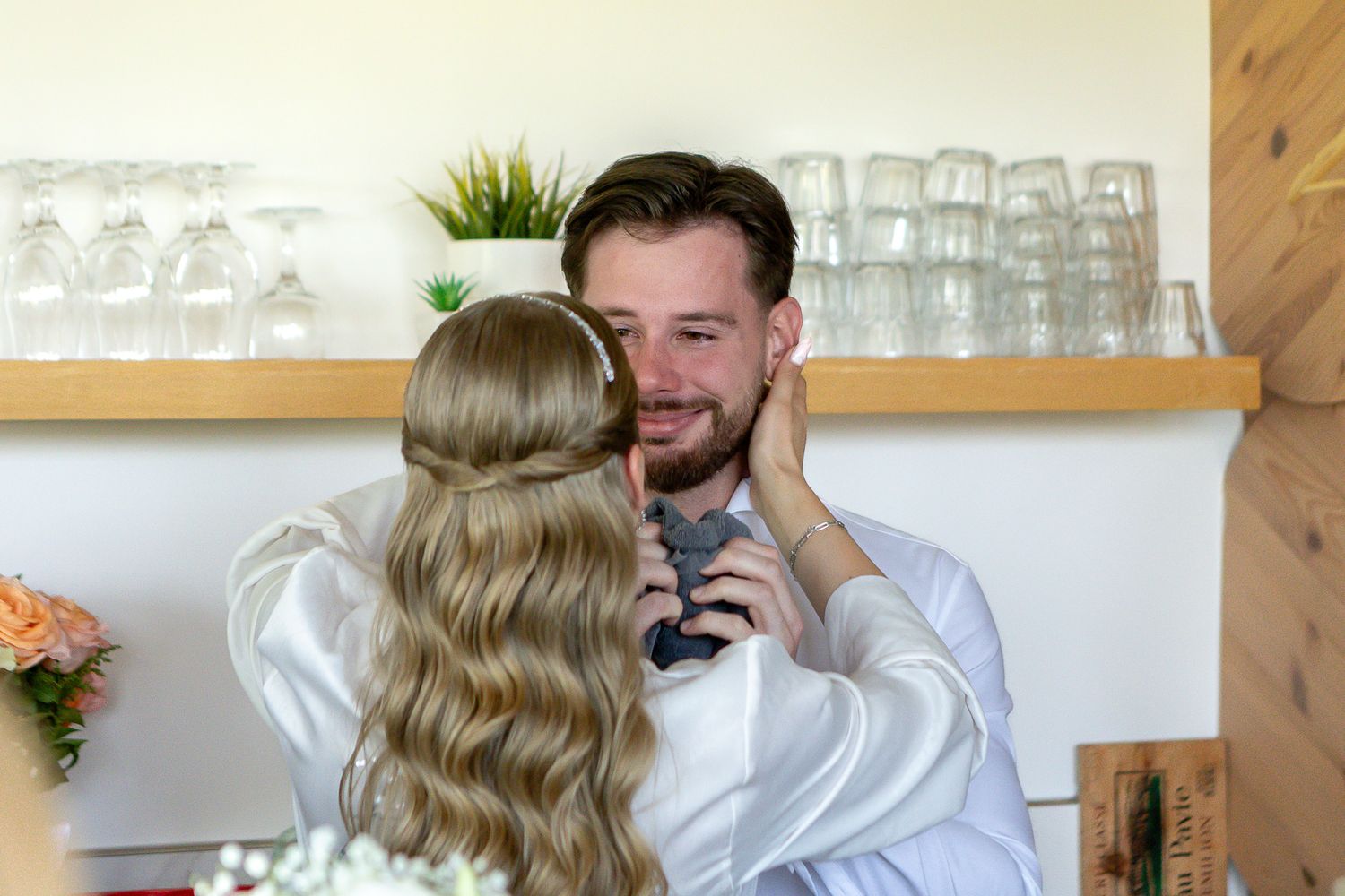 A couple in elegant white wedding attire share a tender moment in a sunlit room.