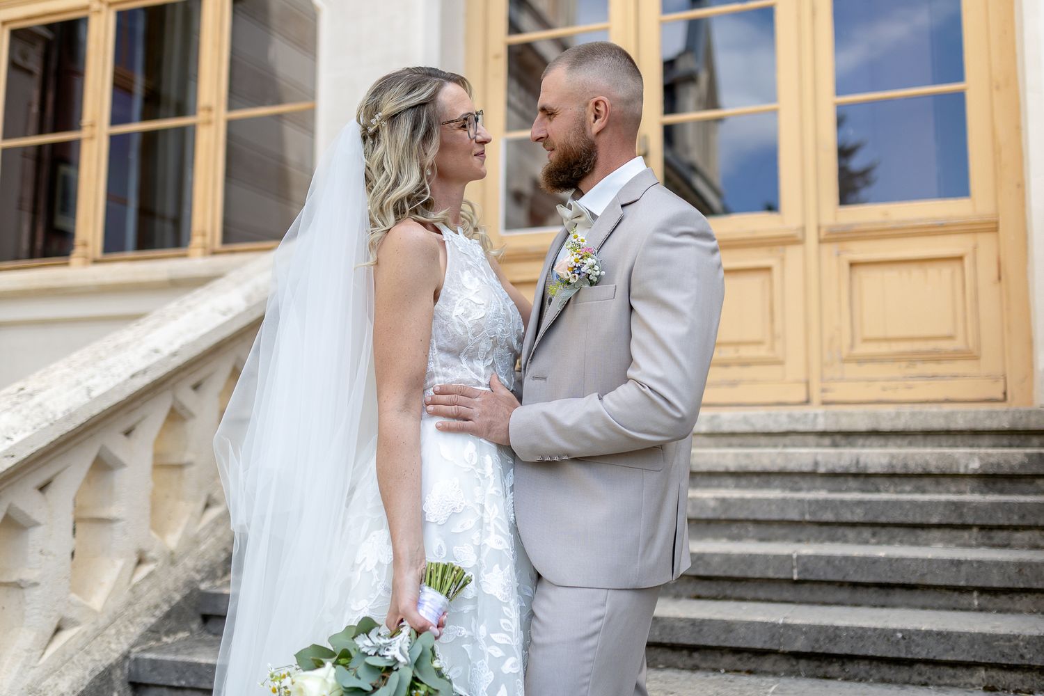 A bride and groom share an intimate moment on elegant stone steps outside a historic building with wooden windows.