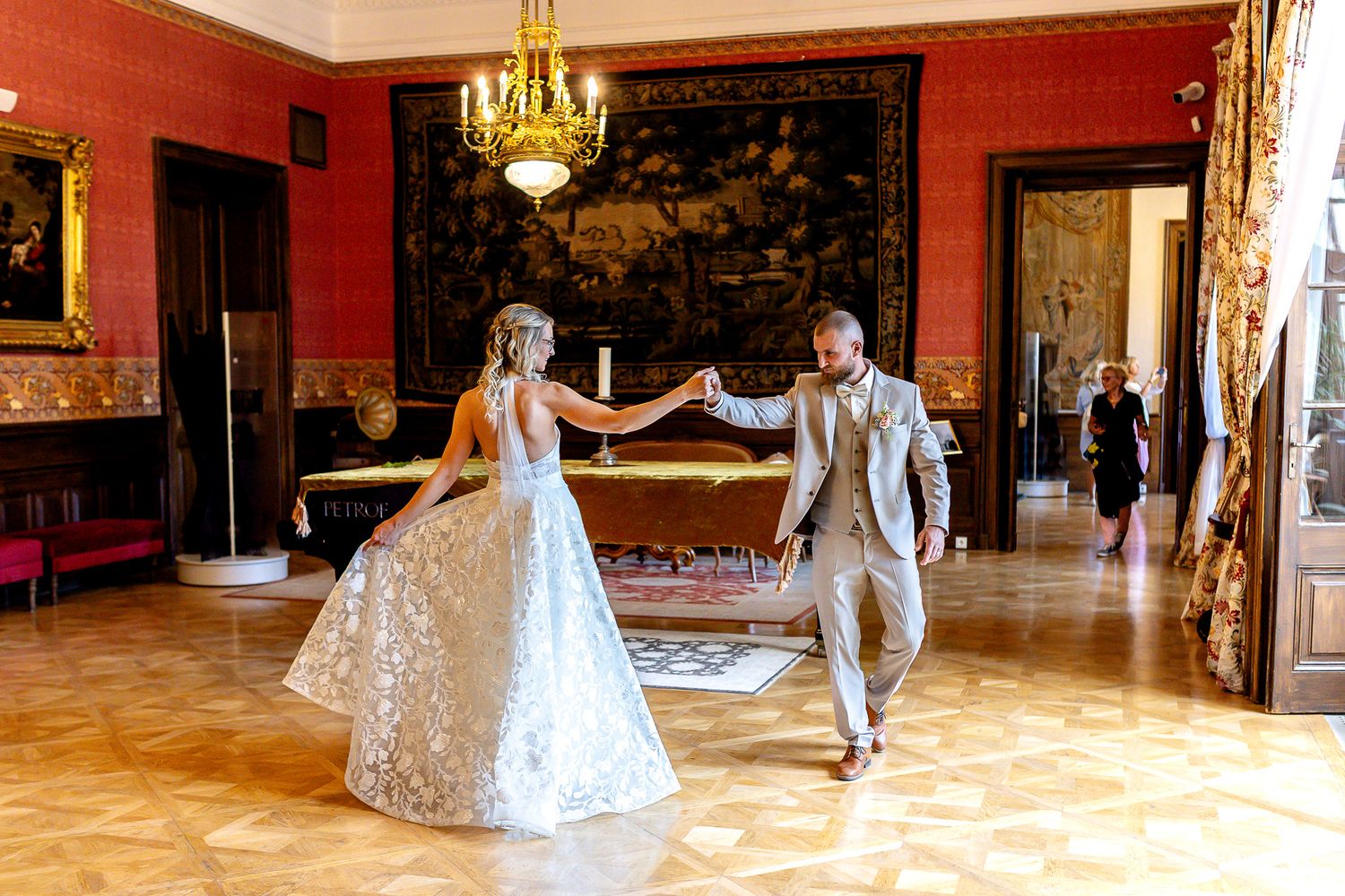 Wedding couple dances in an elegant ballroom with ornate chandeliers and red walls during their first dance celebration.