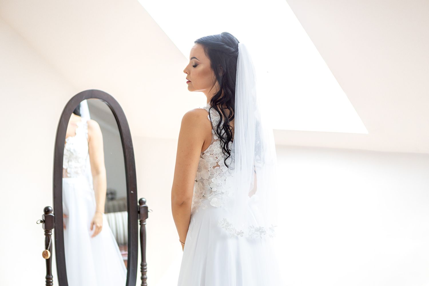 A bride in a white lace wedding dress stands elegantly against a light background.