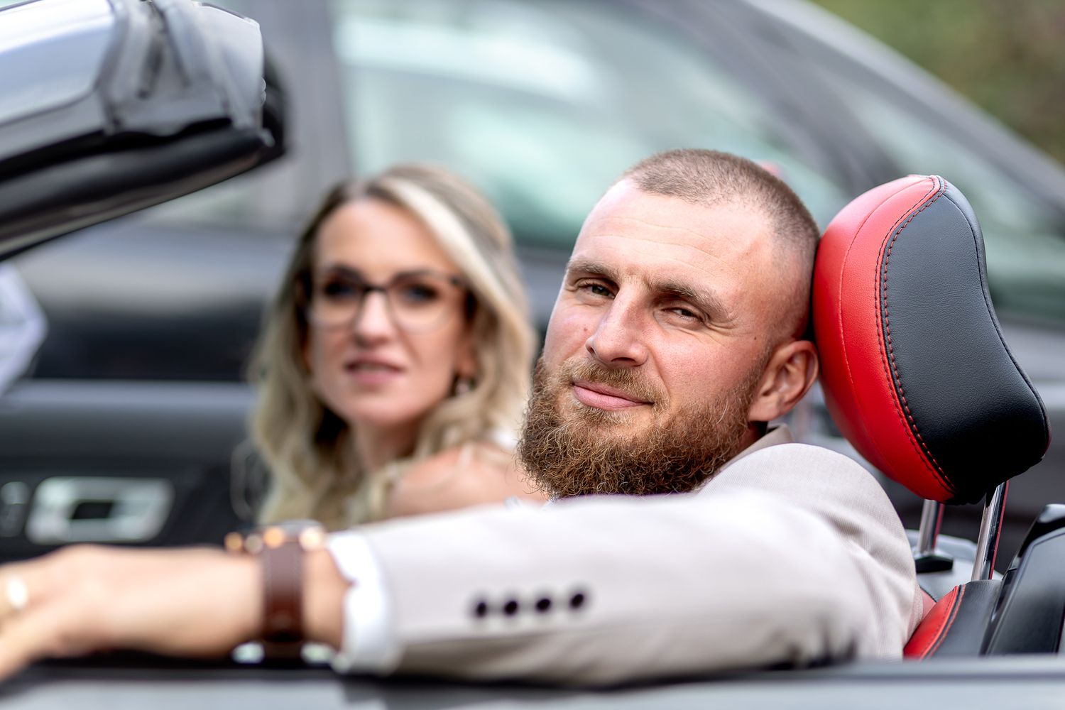 Two people sitting in a luxury convertible car with red leather seats and a casual relaxed atmosphere.