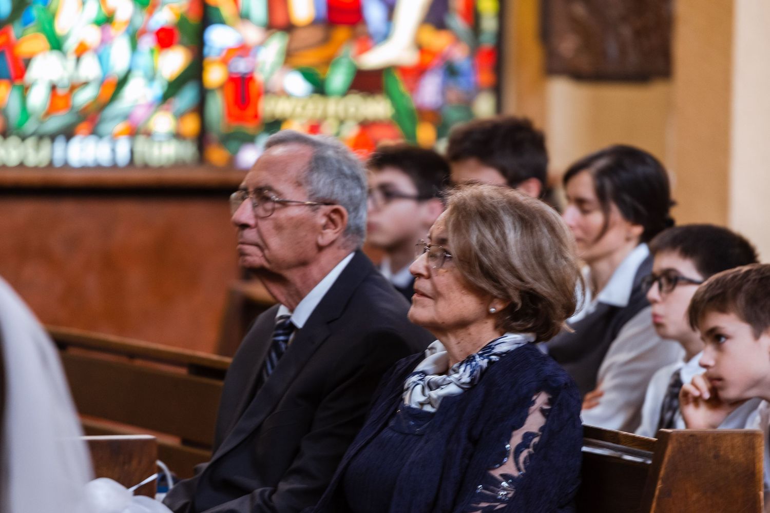 Guests seated in wooden church pews during a ceremony with colorful stained glass windows visible in the background.