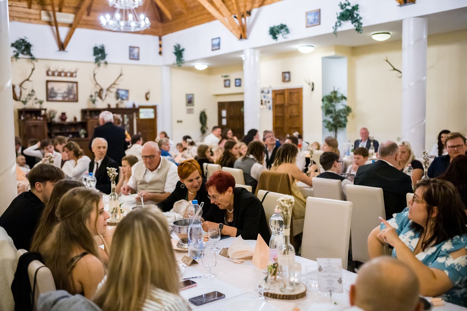 Wedding reception dining room filled with guests seated at long tables in an elegant white-walled venue.