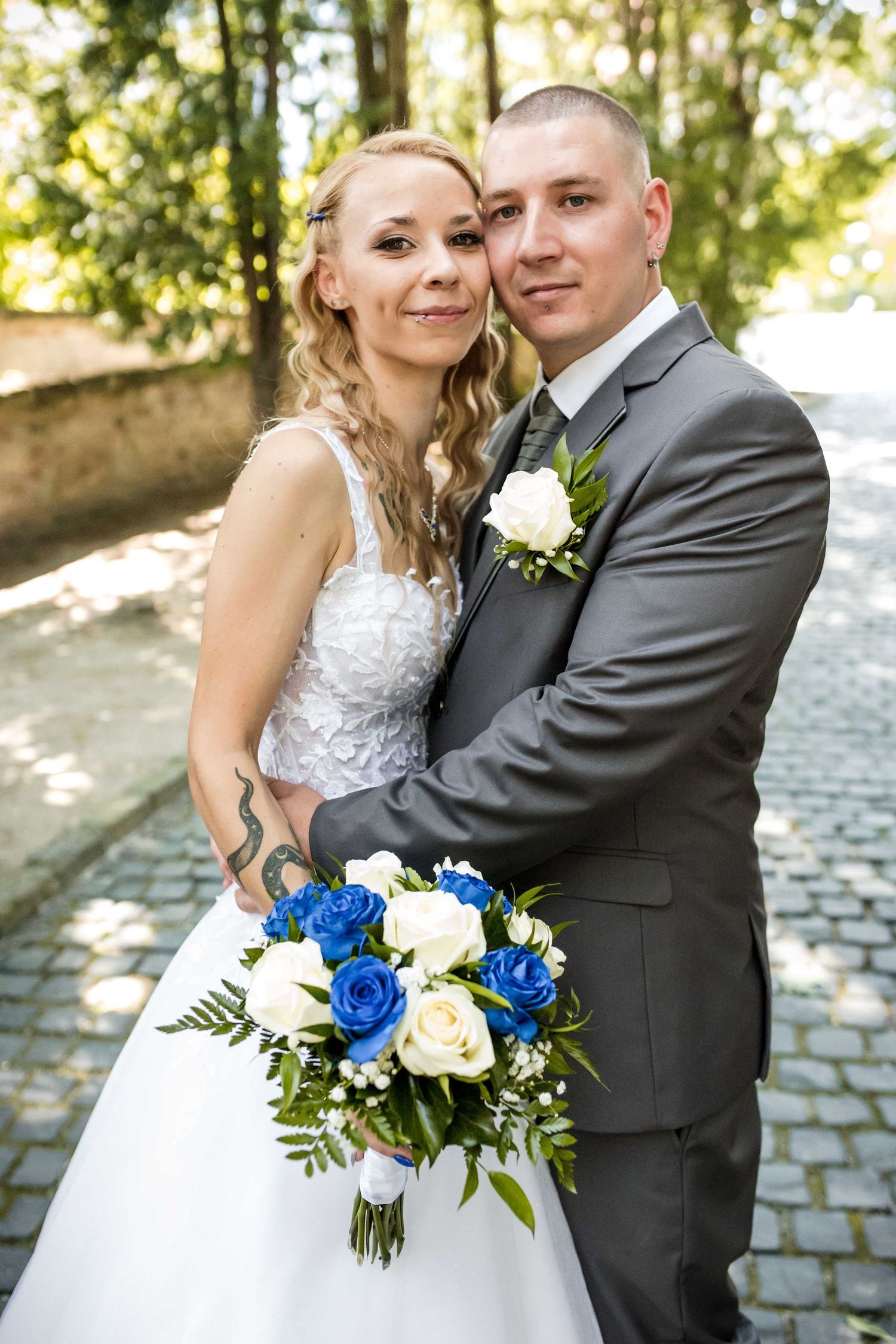 Bride and groom pose outdoors with sunlit trees in background, featuring blue and white wedding bouquet.