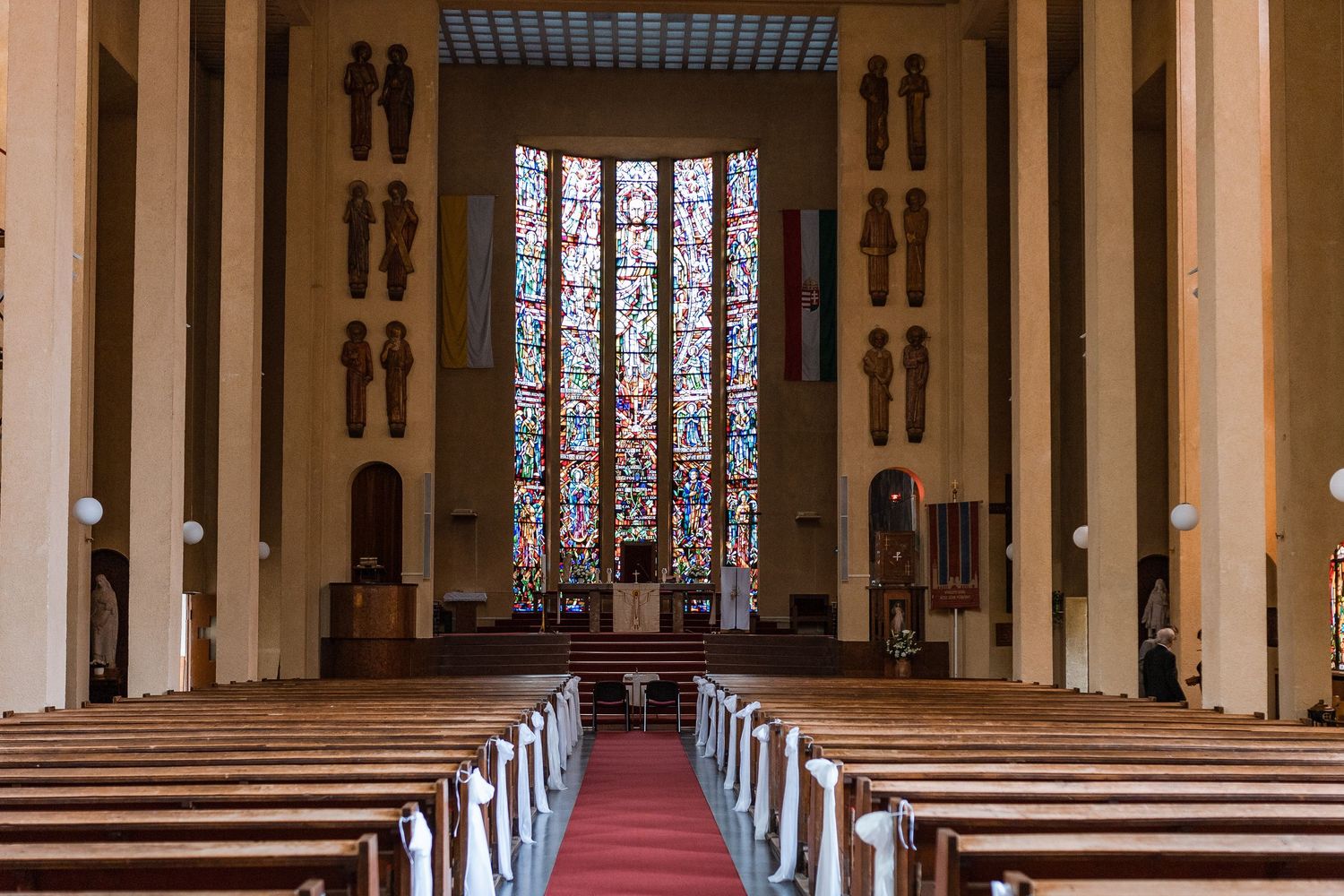 Interior view of a modern church with rows of wooden pews and tall stained glass windows along the walls.