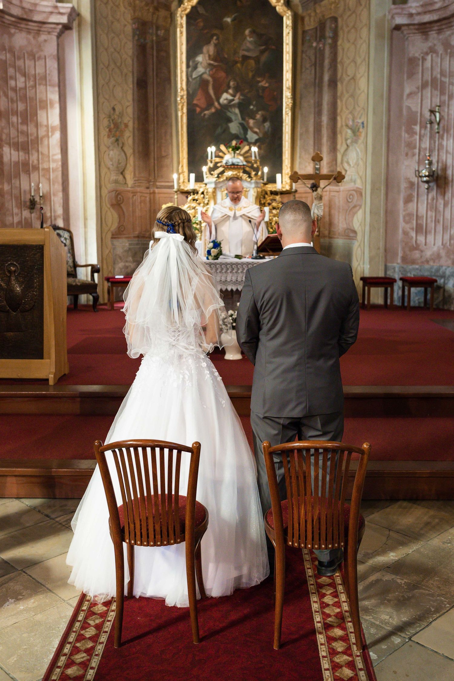 A couple sits on wooden chairs in an ornate church interior with a red carpet runner and gilded decorative elements.