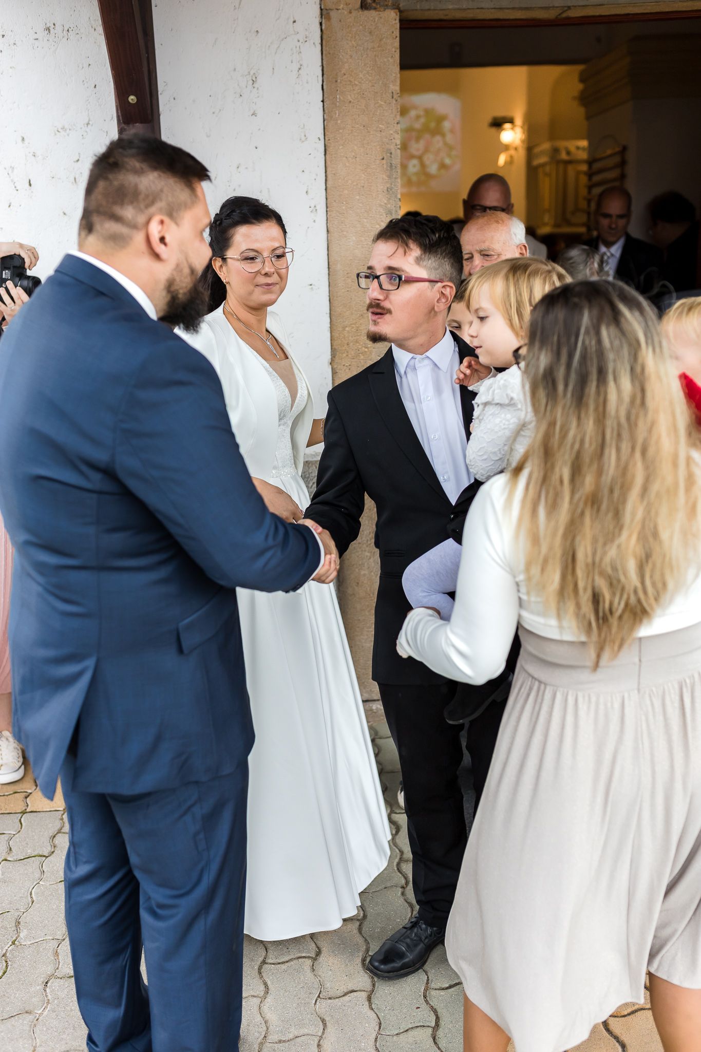 Wedding guests in formal attire gather and mingle at an elegant venue with white stone walls.