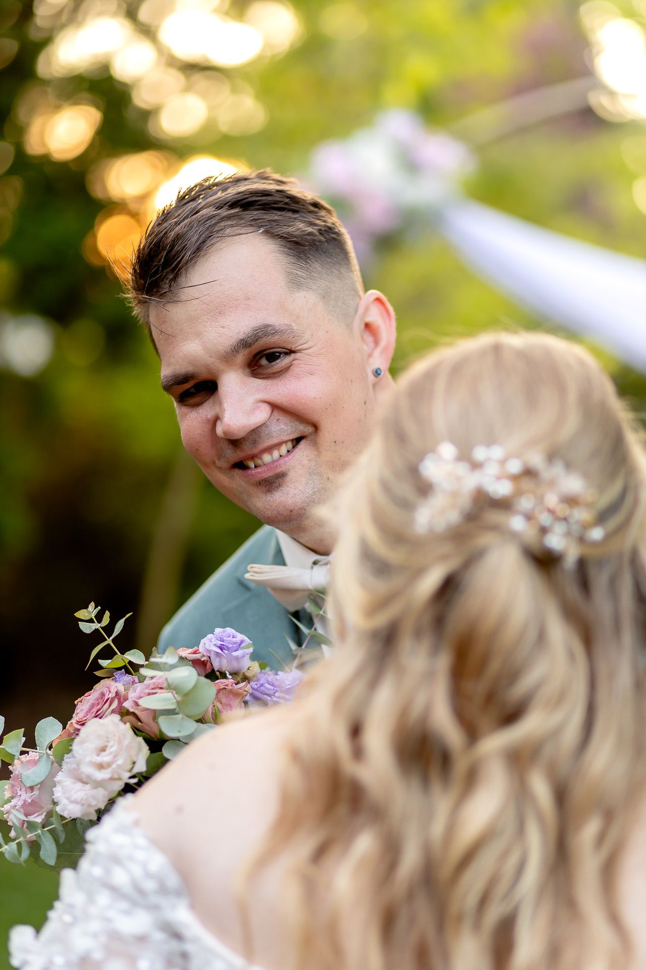 A wedding couple shares a tender moment outdoors with soft bokeh lighting and pastel floral bouquet details.