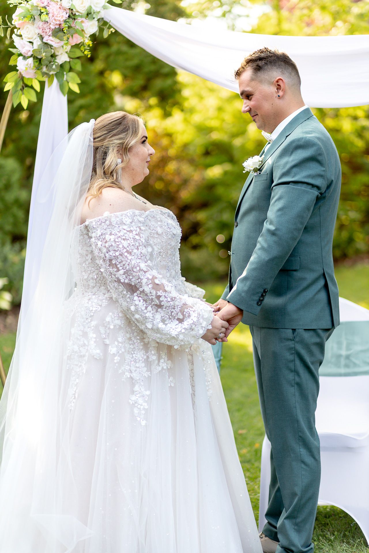 A couple shares a romantic first look moment at their outdoor wedding with white drapery and greenery in the background.