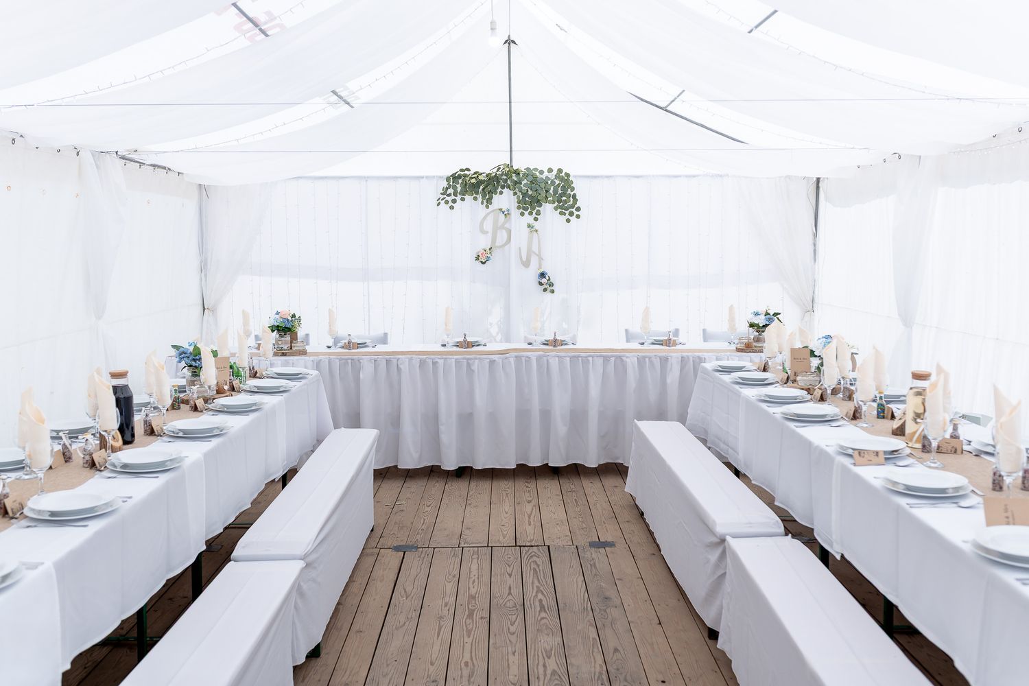 A white tent reception setup with elegant table settings and wooden flooring for an outdoor wedding celebration.