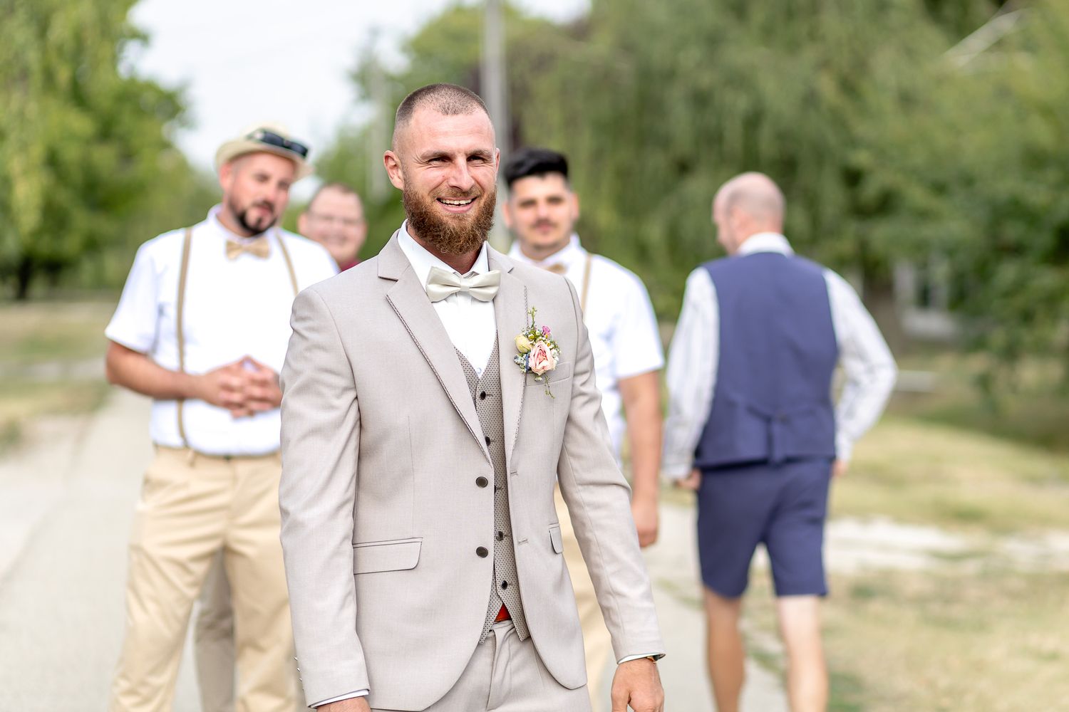 A wedding party in beige and gray attire walks together on a path outdoors during a summer celebration.