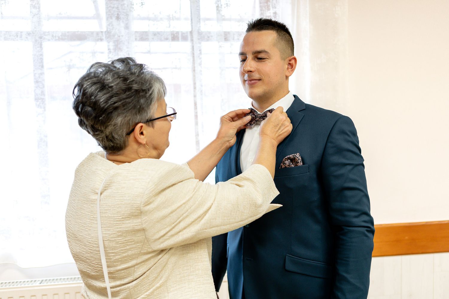 Two people in business attire stand together while one adjusts the other's collar in an office setting.