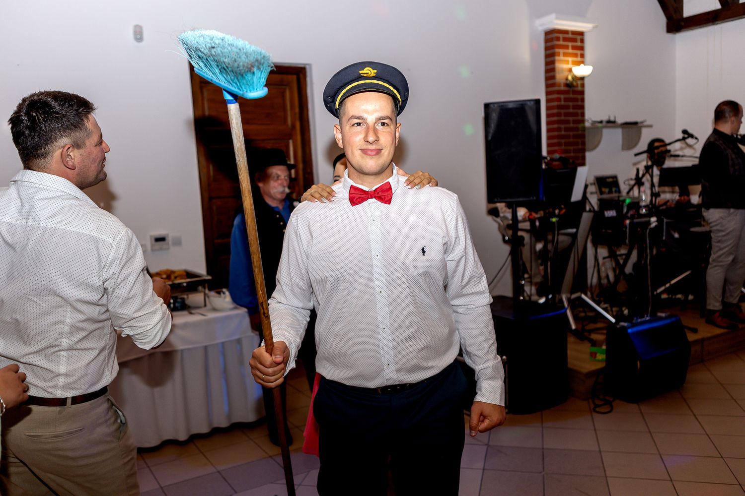 Someone in a white dress shirt, bow tie and cap holds a blue mop while posing at an indoor event.