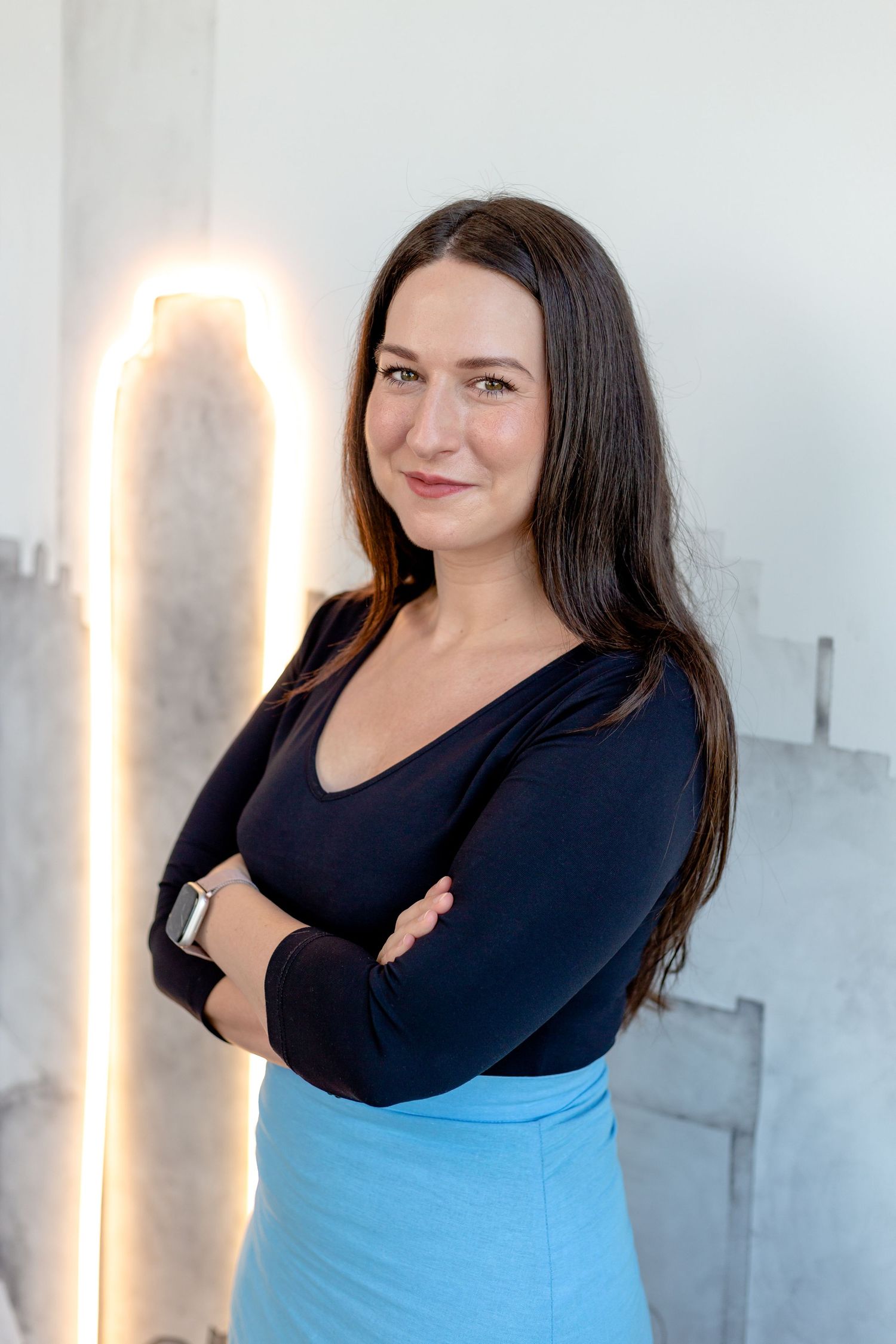 A person in a black top and light blue bottom poses against a white wall with illuminated decor.