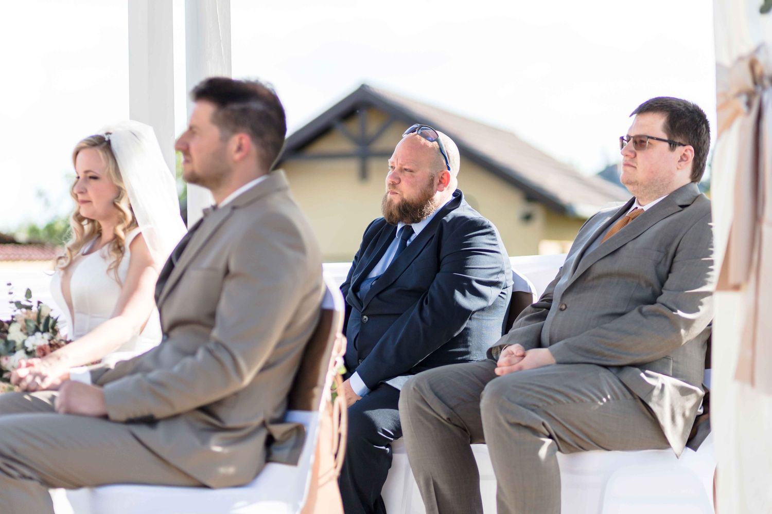 Wedding party members in formal attire laugh together while seated in a row during an outdoor celebration.