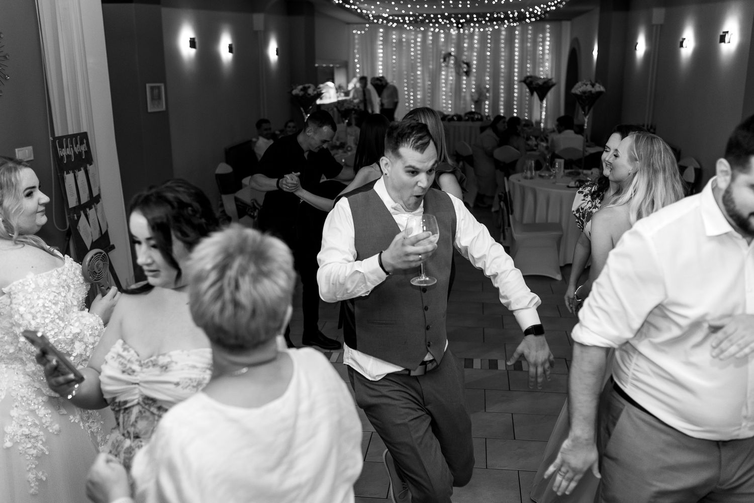 Black and white photo of a wedding reception with string lights overhead and guests dancing and mingling.
