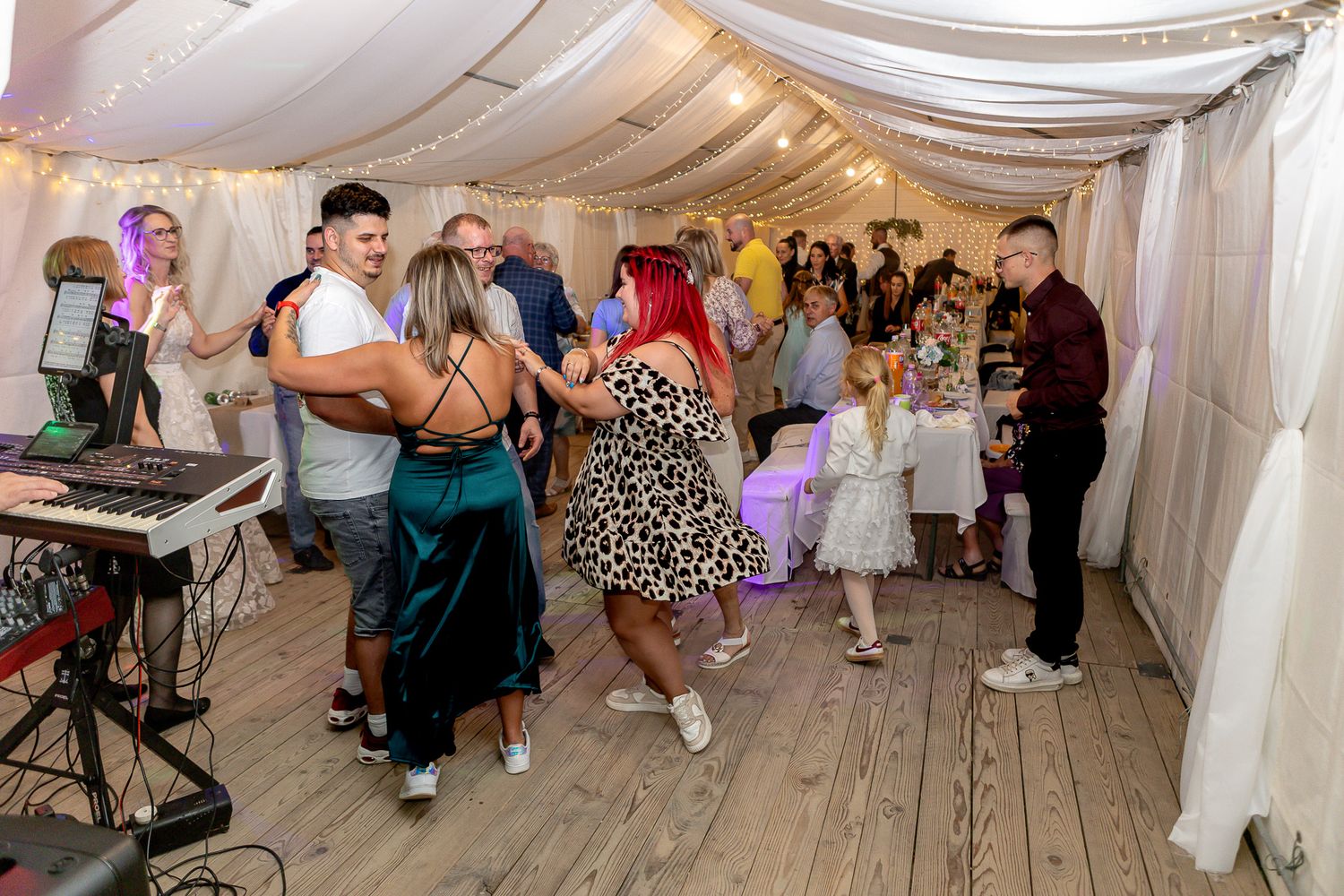 Wedding guests dance and celebrate on a wooden floor under a white tent during a reception party.