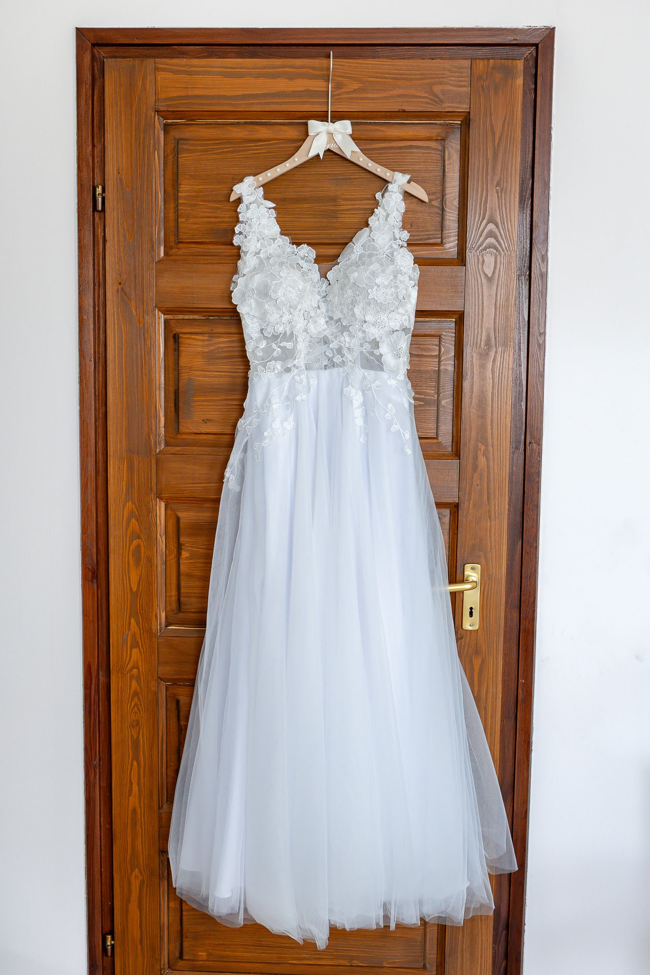 White tulle flower girl dress with lace bodice and delicate straps hangs on wooden door for display.