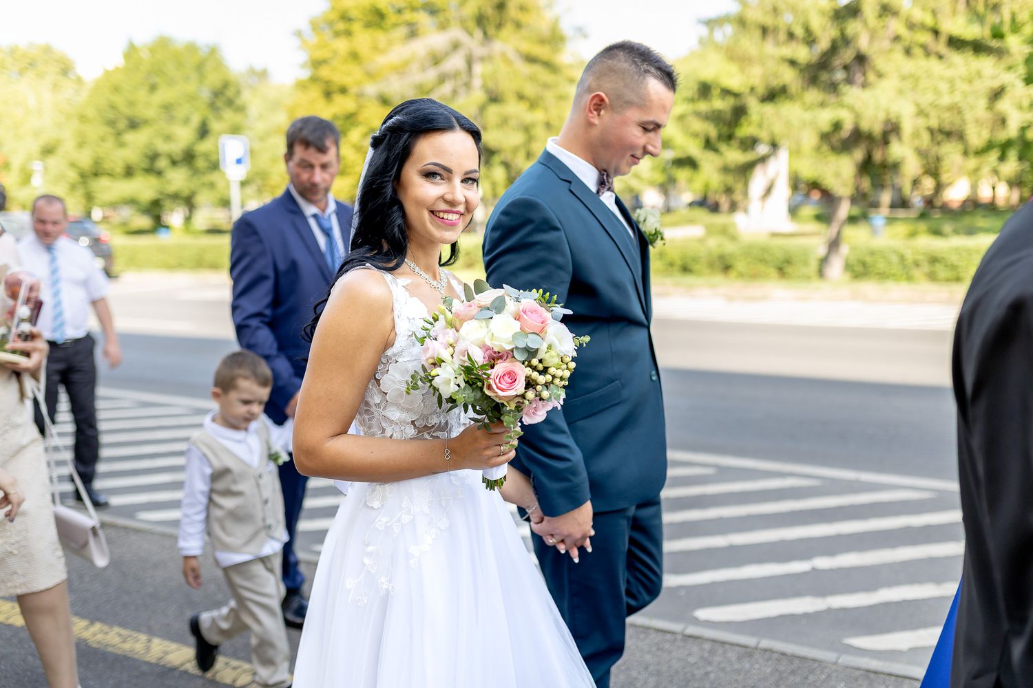 Wedding party members walk and laugh together in a sunny outdoor parking lot on a summer day.