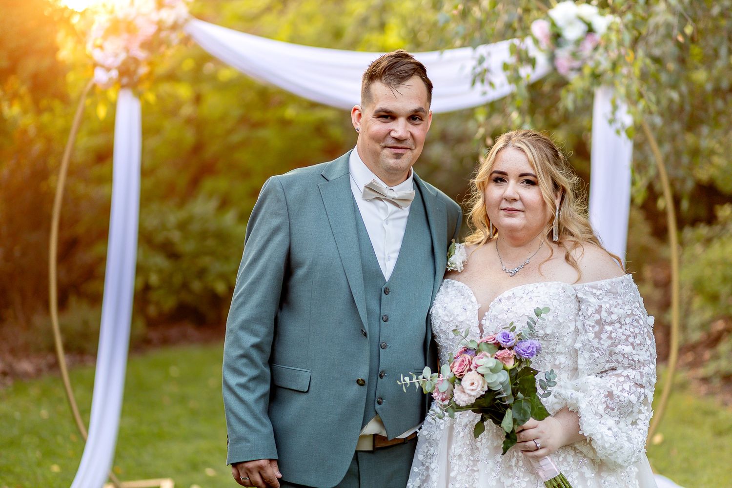 A couple poses outdoors on their wedding day with autumn foliage and golden sunlight in the background.