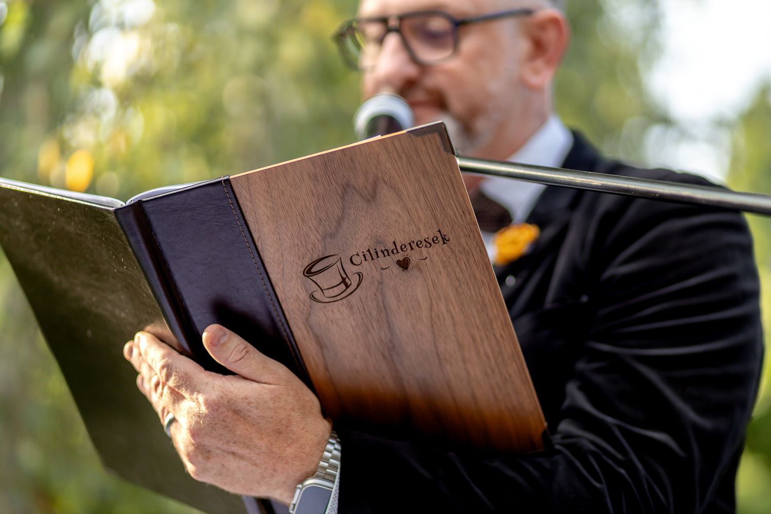 A person in a black suit jacket reads from a brown leather-bound book outdoors with trees in the background.