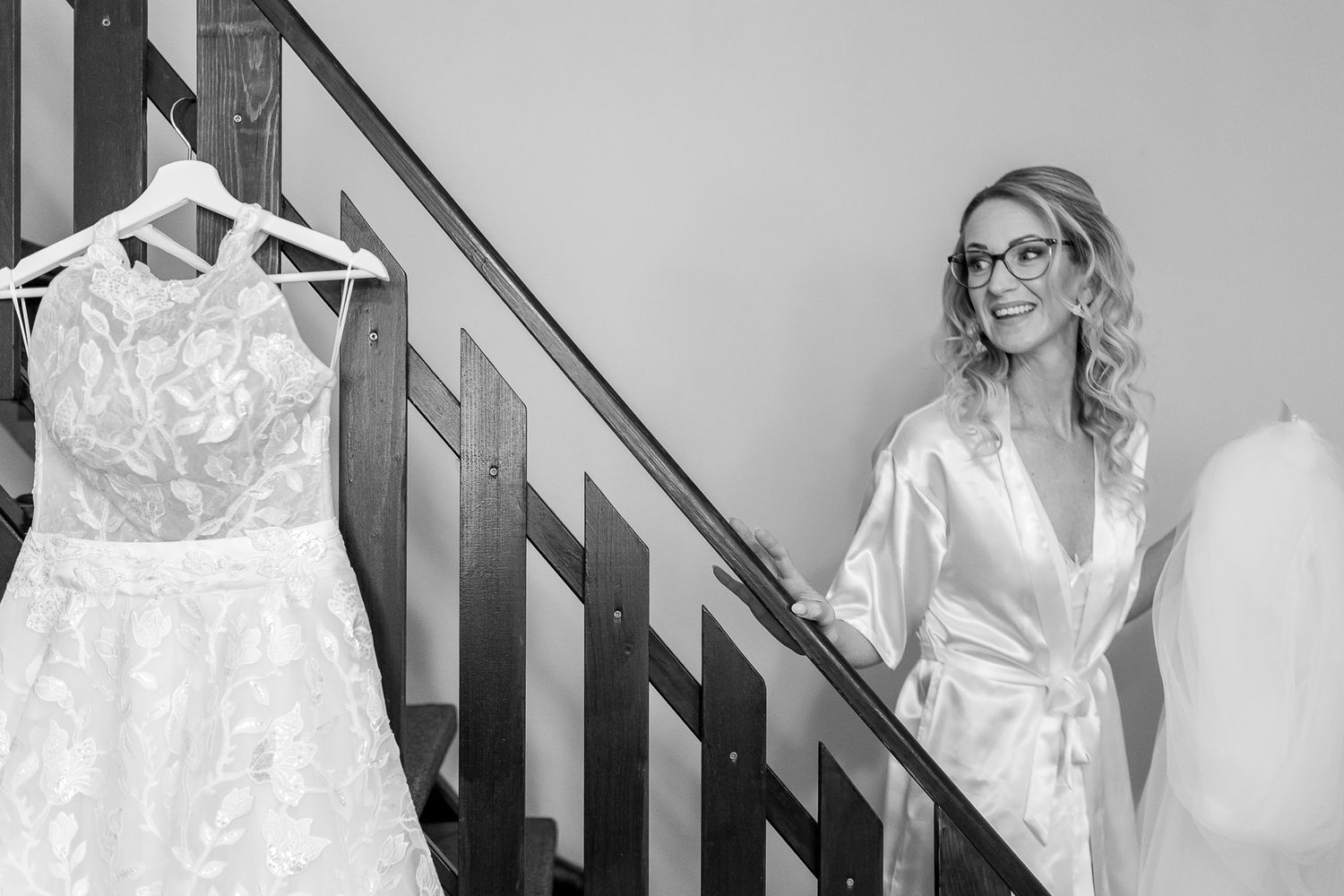 A wedding dress hangs on display while someone in a silk robe stands on a staircase during pre-wedding preparations.