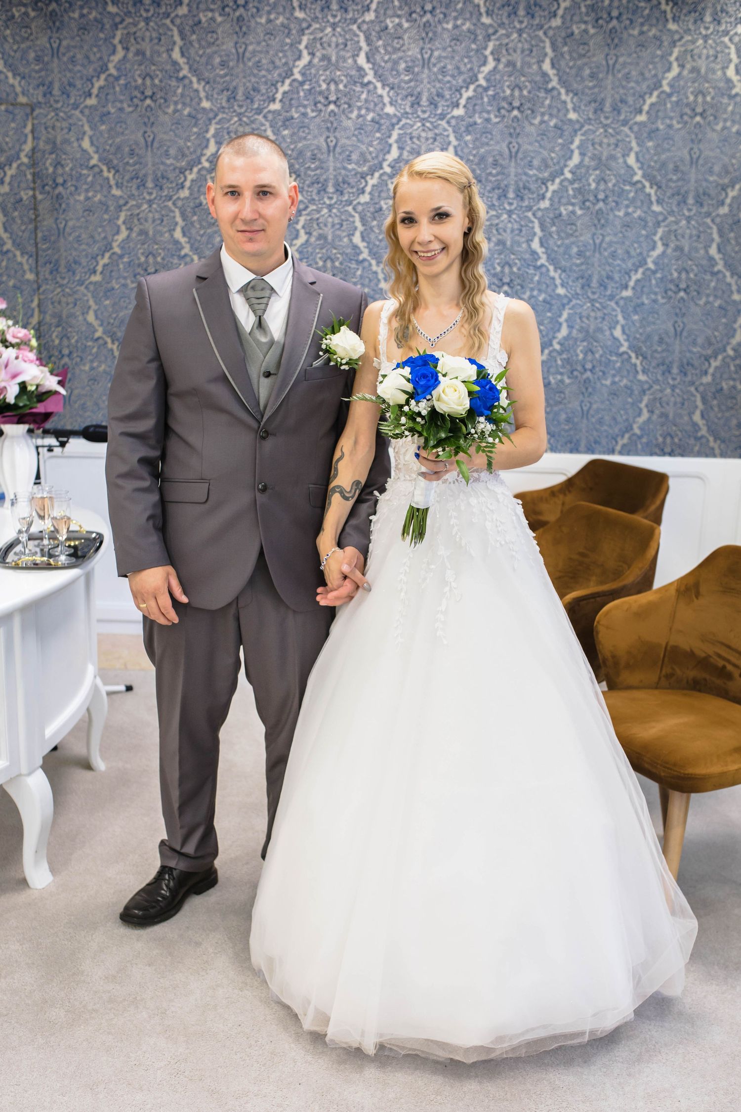 Elegant wedding scene with bride in white gown and groom in grey suit posing against decorative wall.