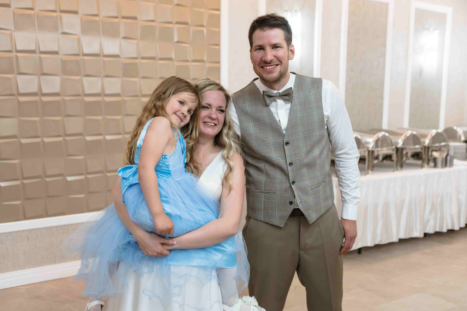 Three people in coordinating blue and gray formal attire pose together at an event venue with decorative wall panels.