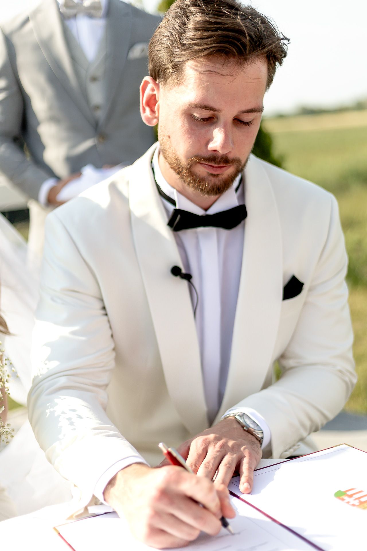 Groom in white tuxedo signs marriage documents at outdoor ceremony.