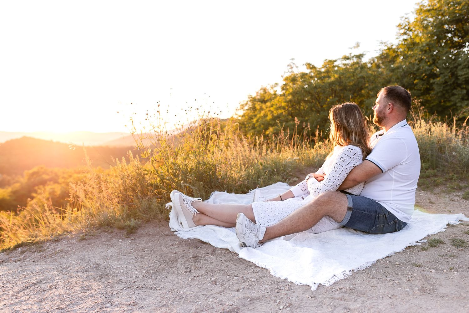 A couple sits on a white blanket enjoying a romantic sunset in a scenic mountain setting.