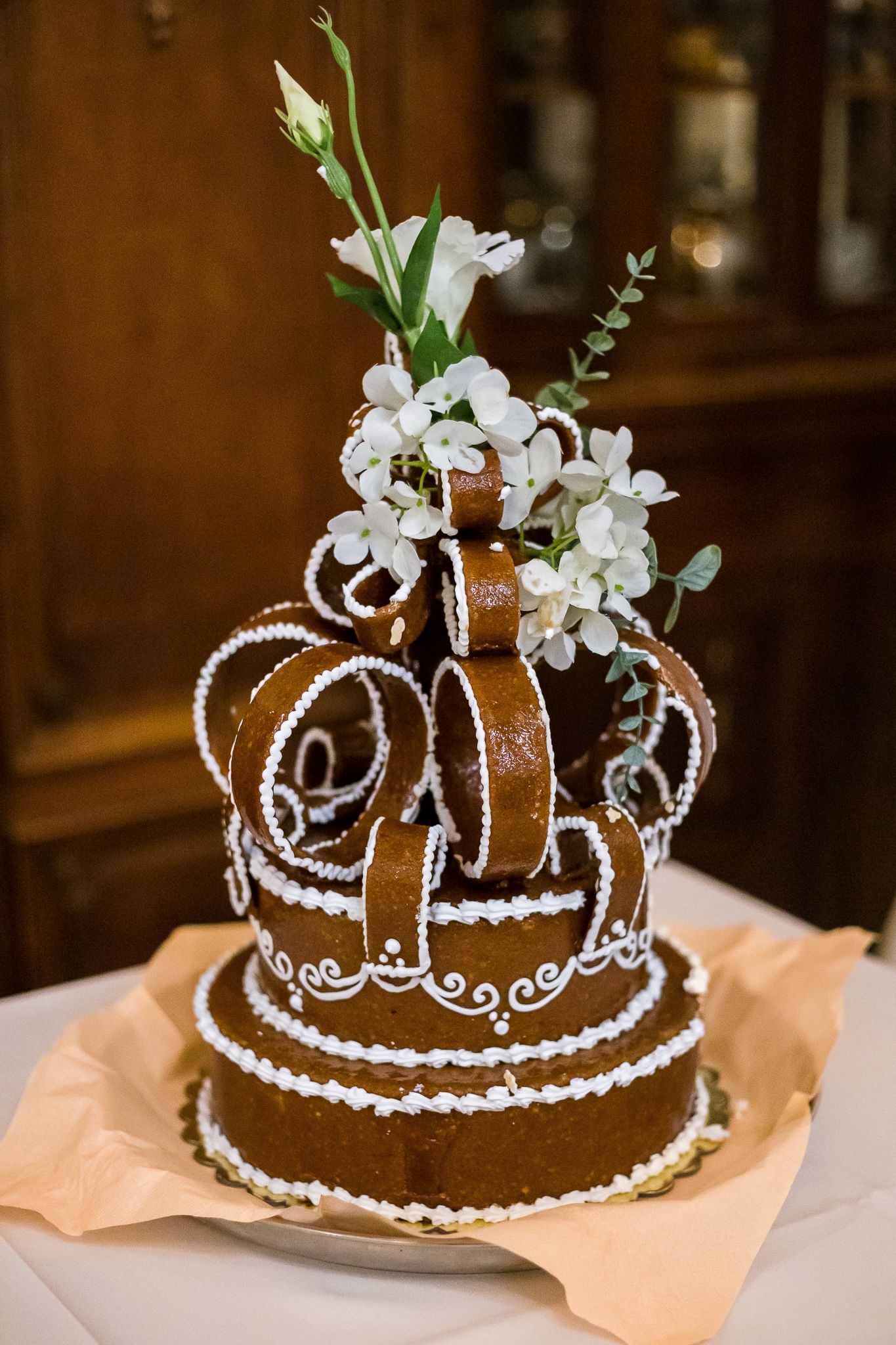 A chocolate brown crown-shaped cake decorated with white icing pearls and floral accents sits on peach-colored fabric.