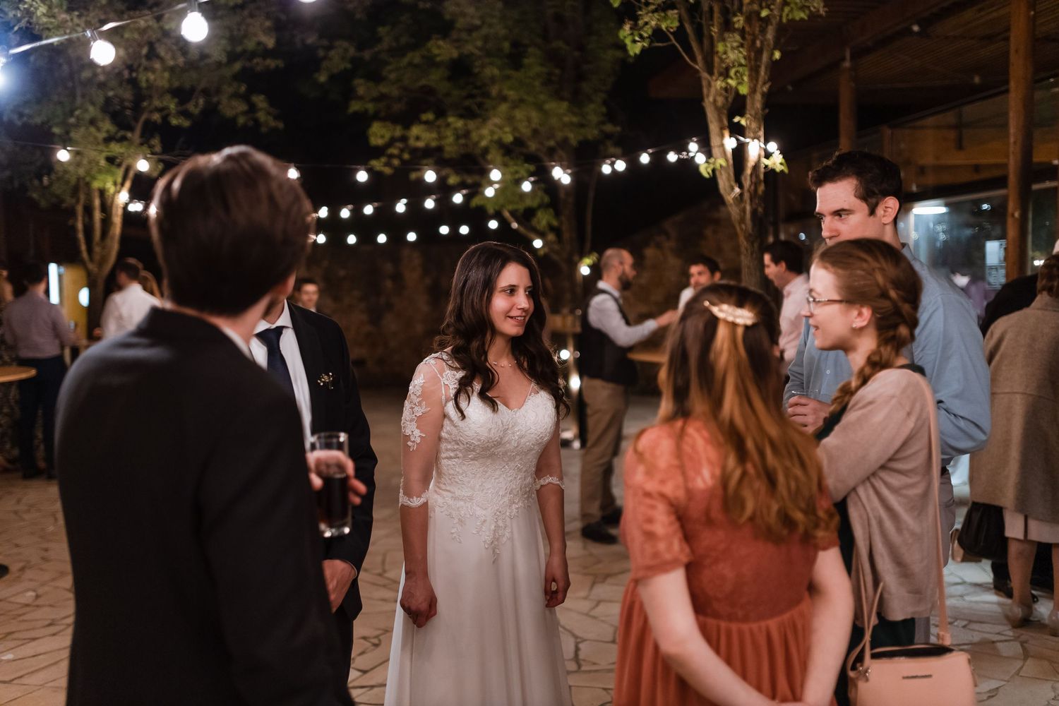 Wedding guests dance and celebrate under glowing string lights at an outdoor evening reception.