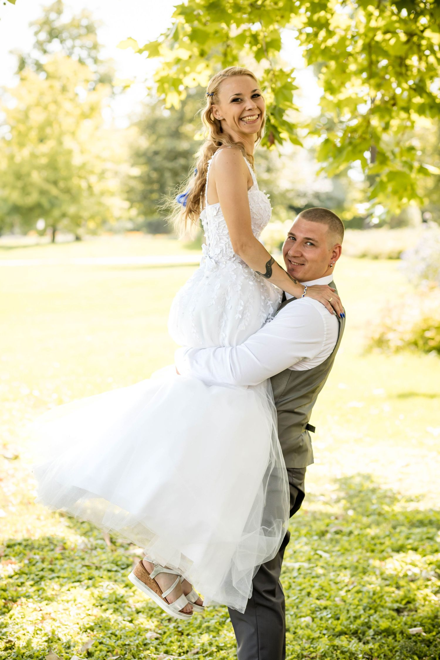 A couple in wedding attire embraces joyfully in a sun-dappled outdoor setting with green foliage in the background.