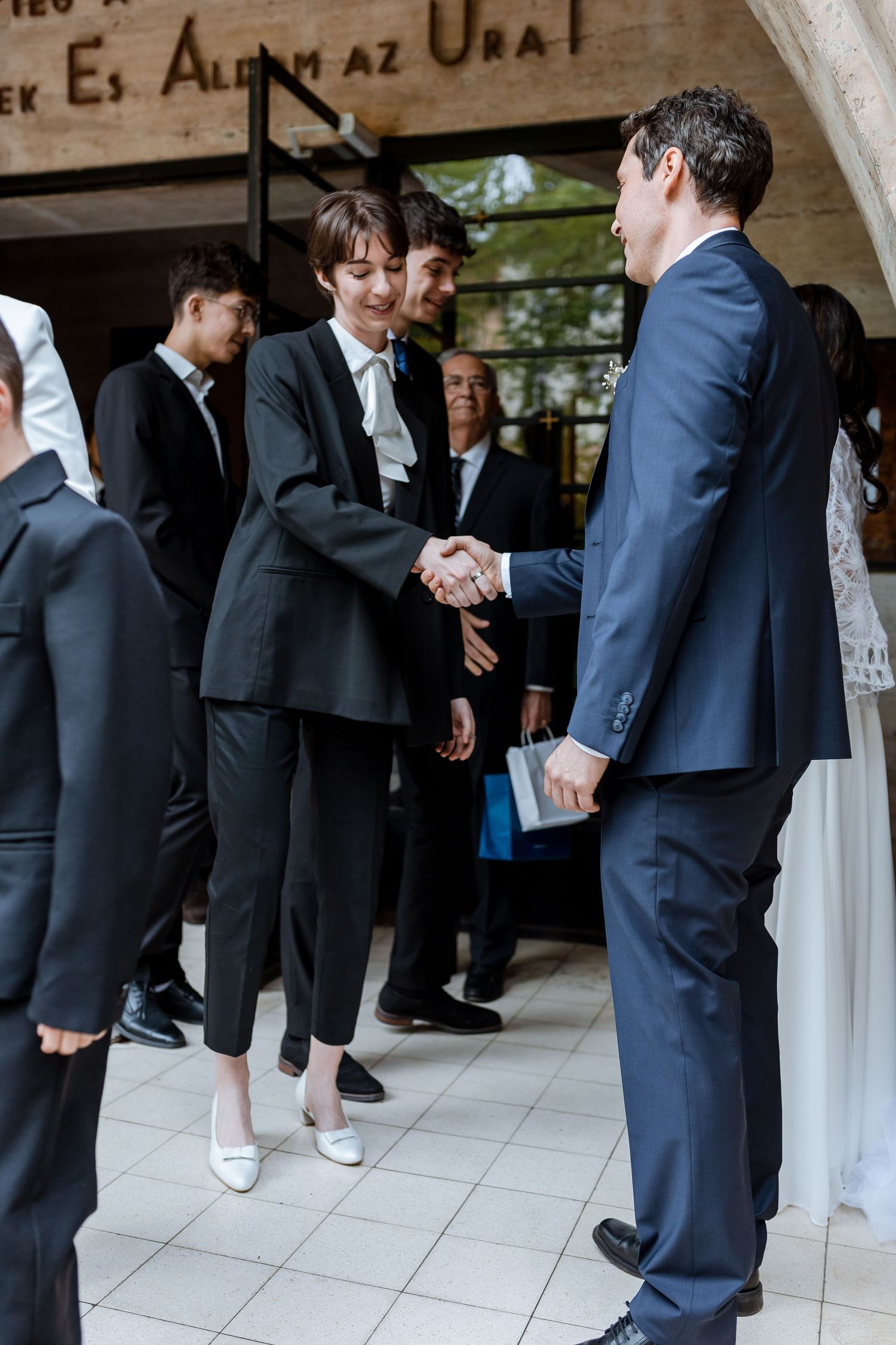 Wedding guests in formal attire greet and mingle in an outdoor covered walkway.