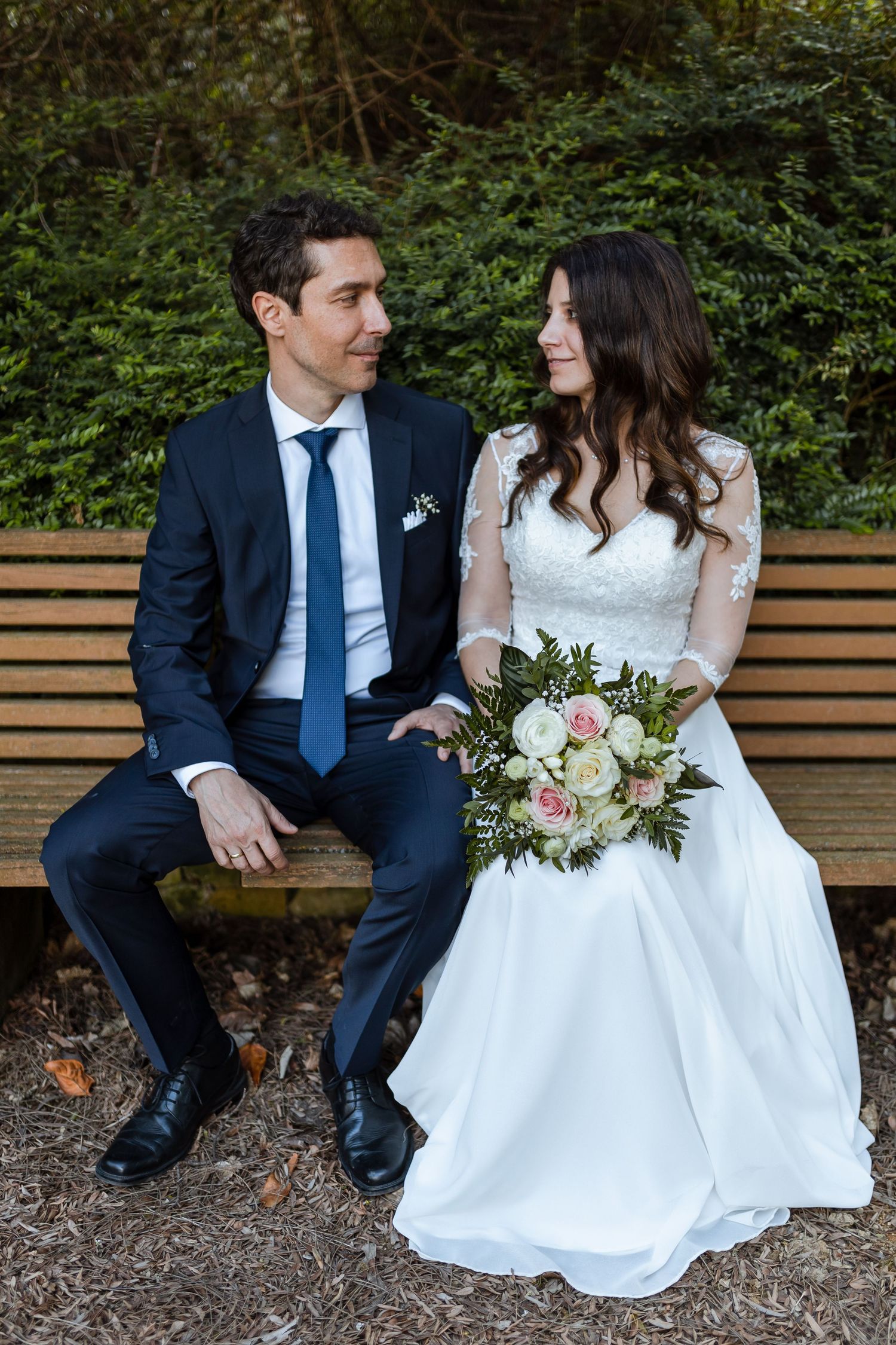 A romantic wedding couple sitting on a wooden bench in a garden setting, sharing intimate moments together.
