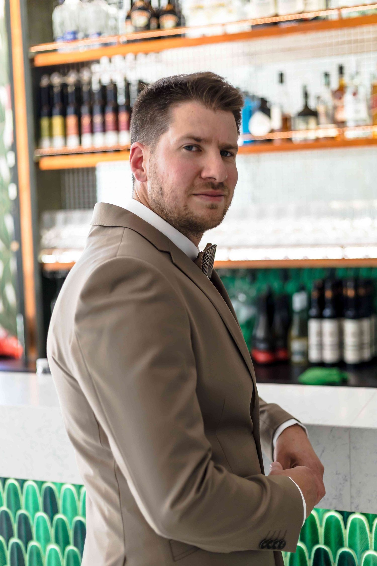 Person in tan suit jacket seated at bar counter with bottles and glassware visible in background.