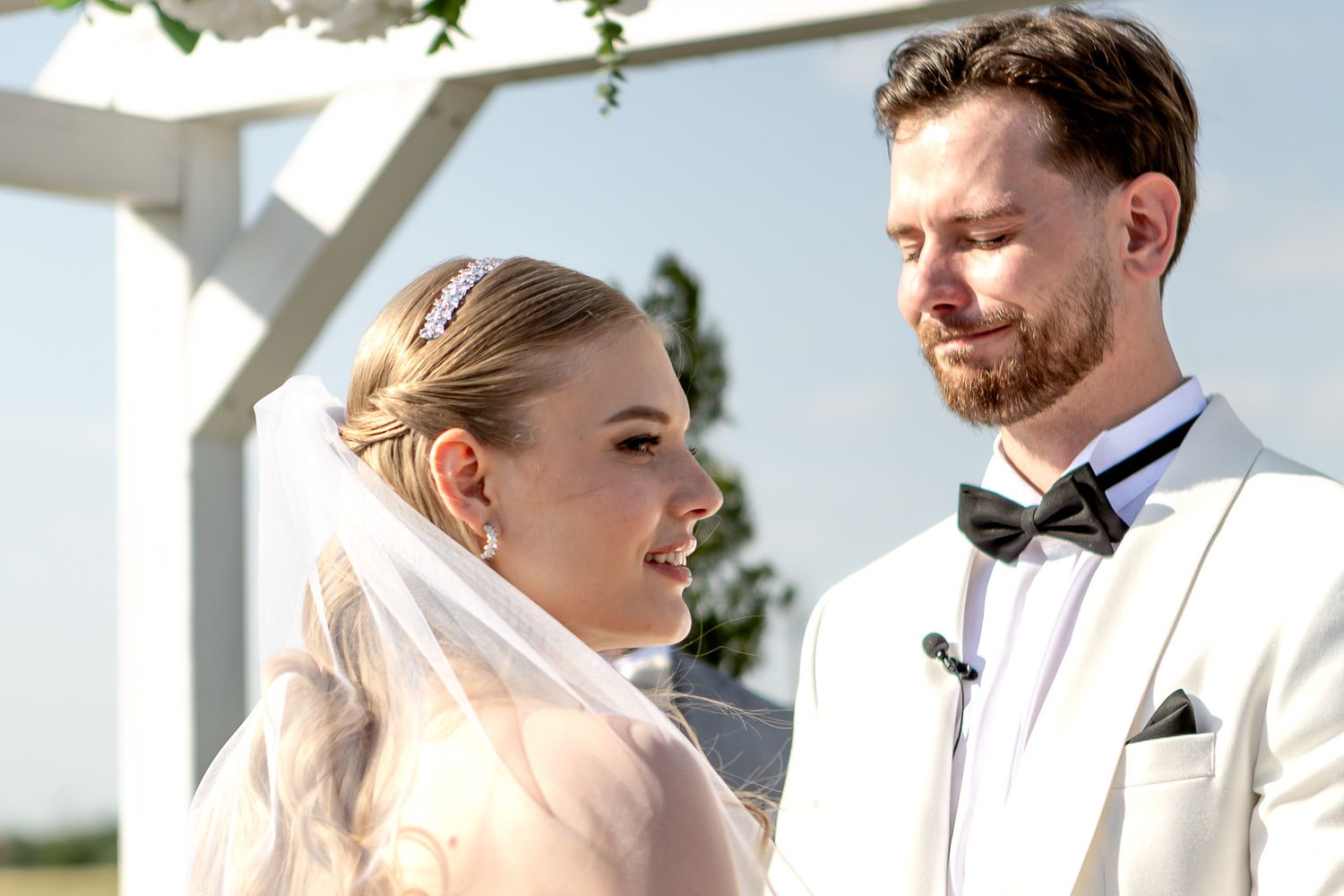Wedding couple exchanges vows during outdoor ceremony under white canopy.
