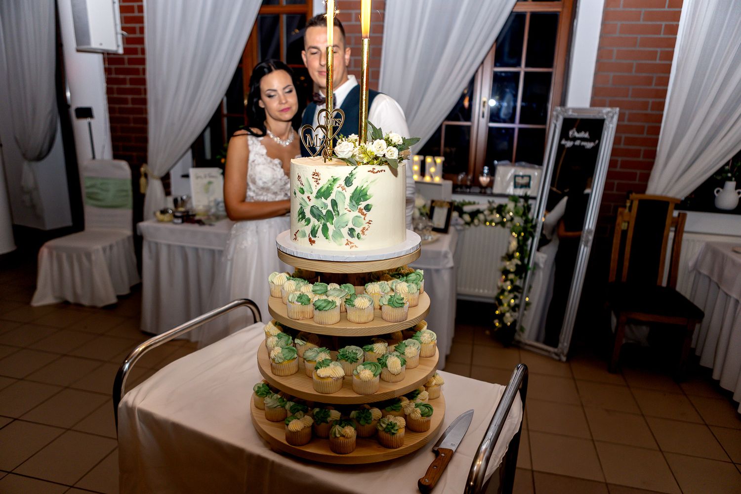 A multi-tiered wedding cake decorated with white frosting and green floral accents displayed at an elegant reception venue.