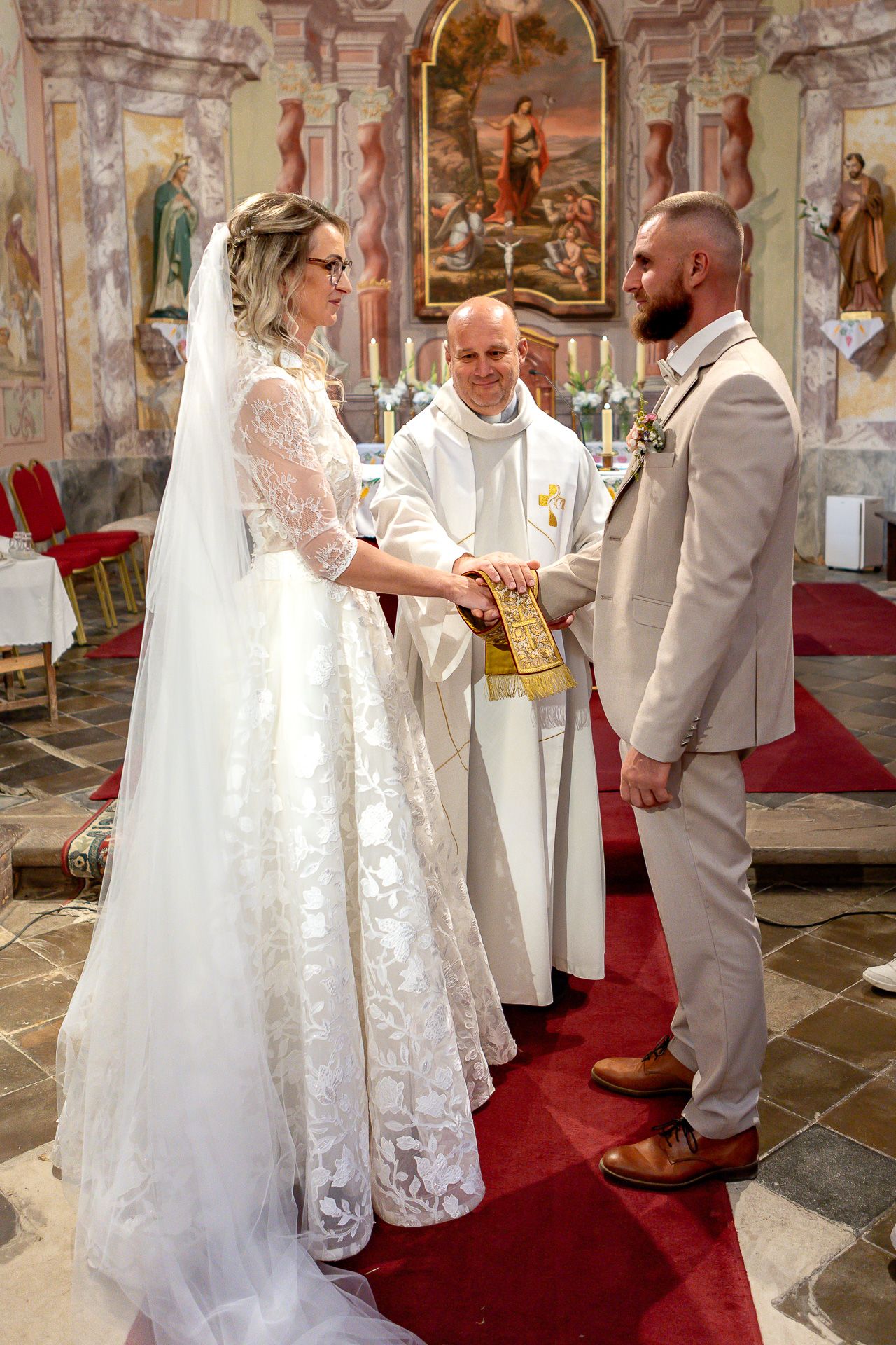 A wedding ceremony taking place in an ornate church with frescoed walls and a red carpet aisle.