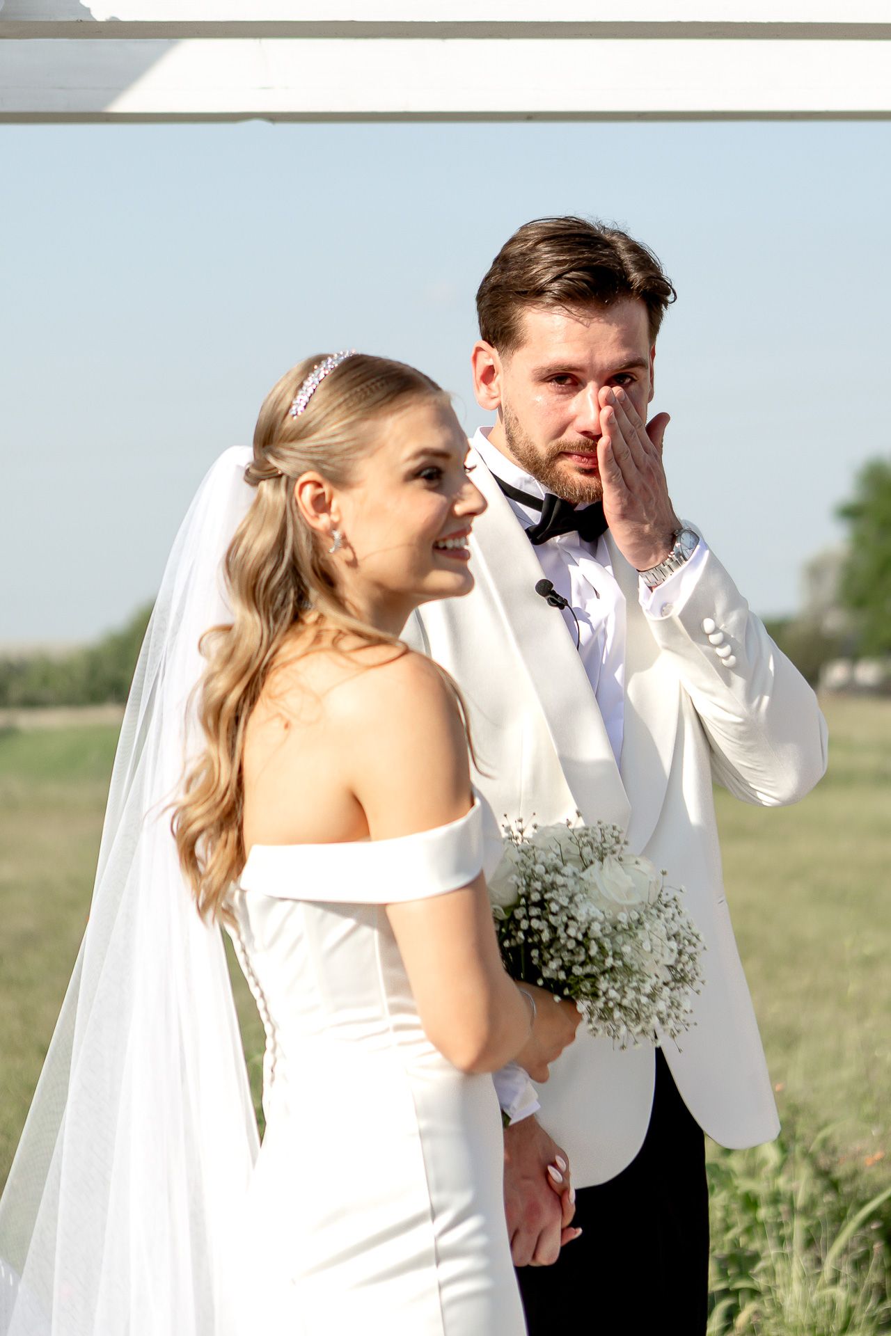 Couple shares an emotional moment during outdoor wedding ceremony with white decor and floral bouquet.