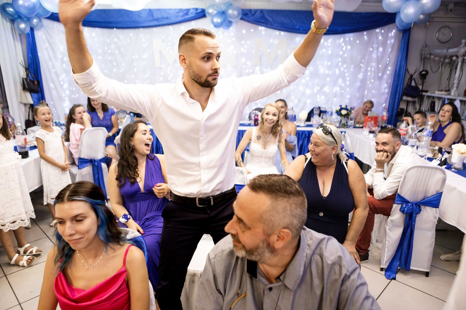 Wedding guests dancing and celebrating with blue decorative fabric draped above at an indoor reception party.