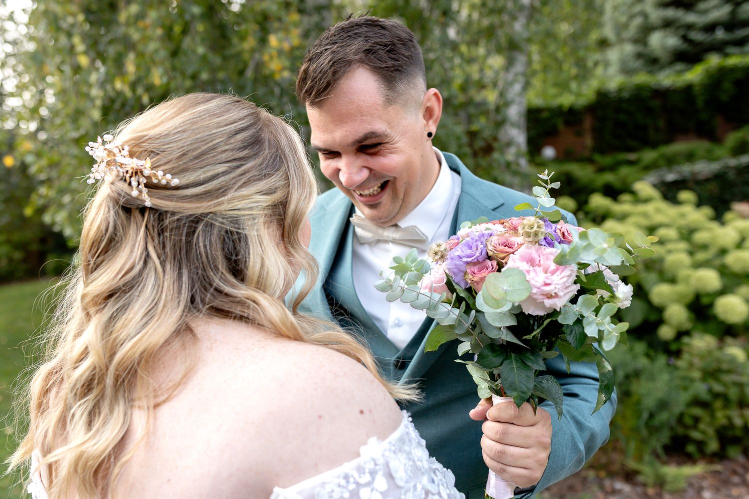 Newlyweds share a tender moment in a lush garden setting with soft pink and white floral bouquet.