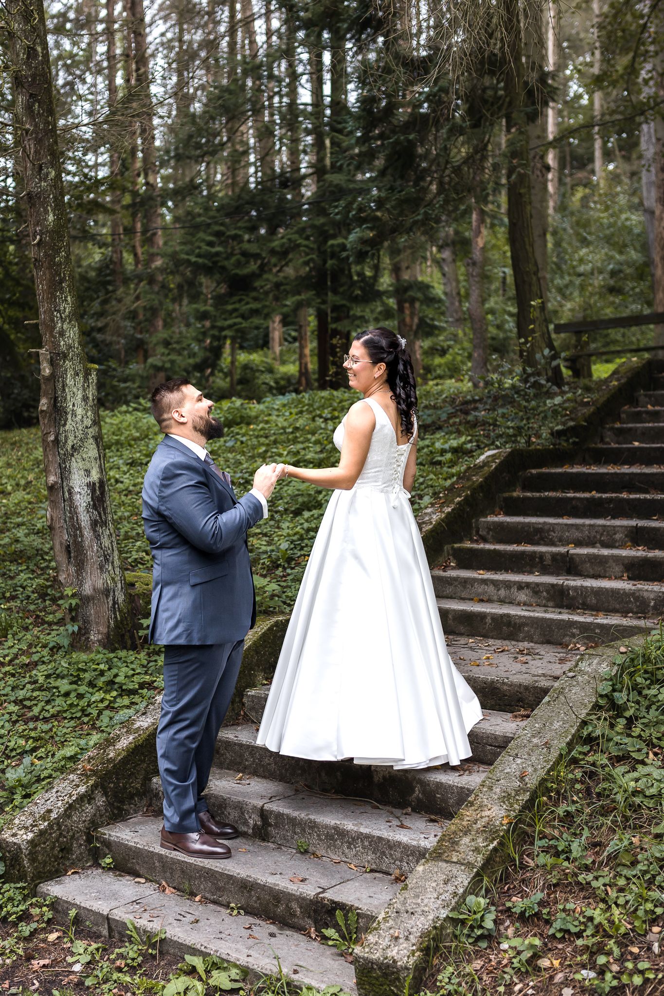 A couple in wedding attire shares a romantic moment on rustic stone steps surrounded by lush forest greenery.