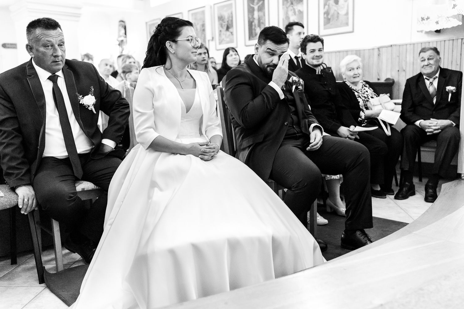 Wedding guests sit in chairs during an indoor ceremony as a bride in a white gown walks past.