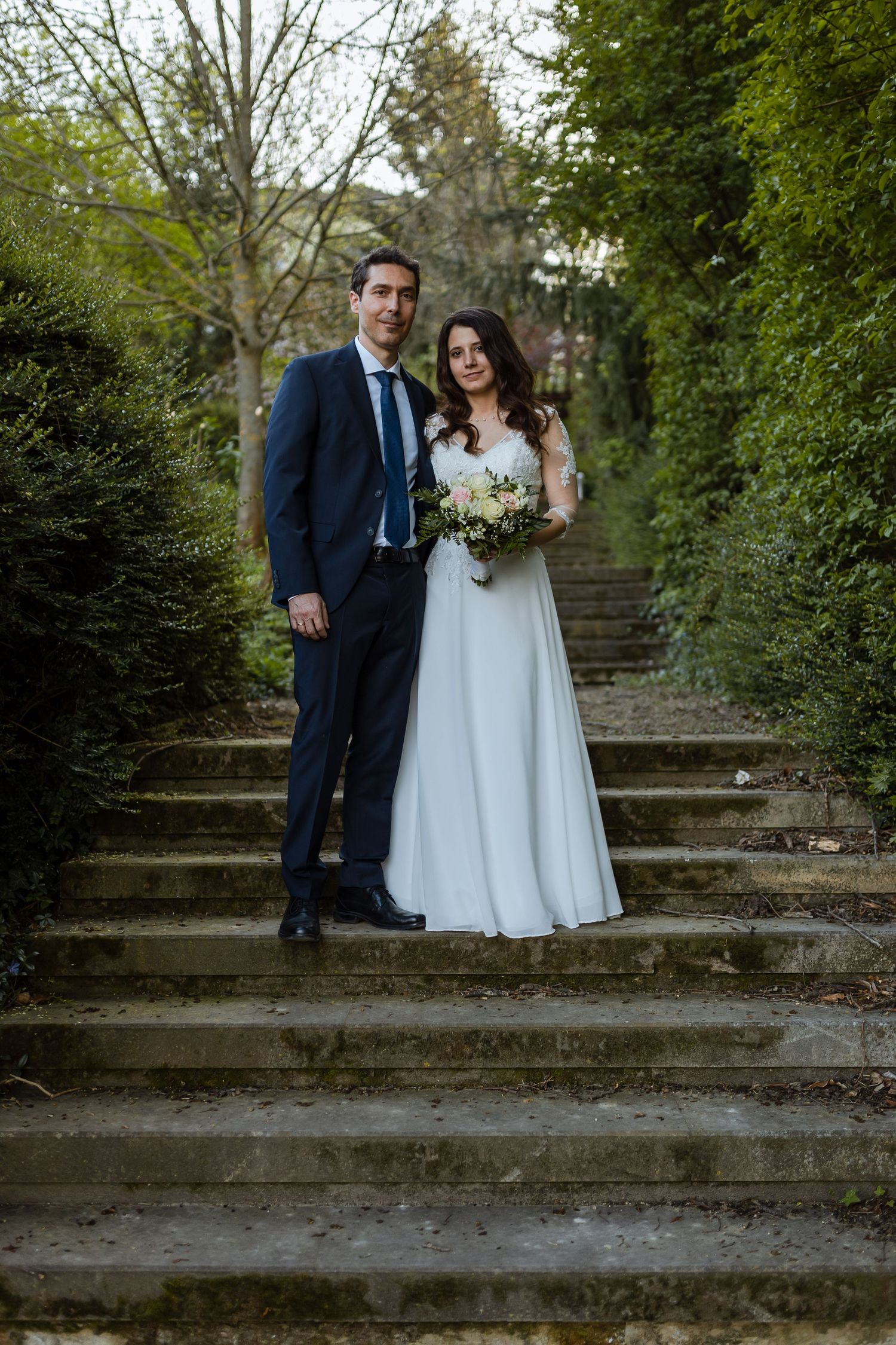 A couple in wedding attire poses on stone steps surrounded by lush greenery in a garden setting.