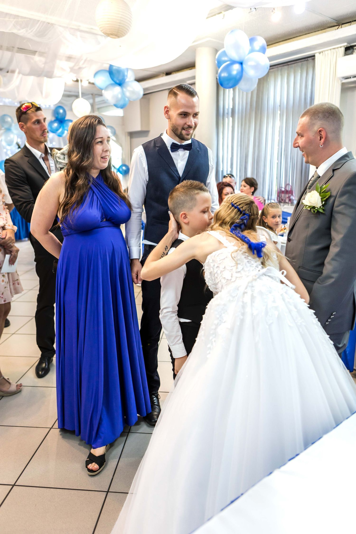 Wedding celebration scene with guests in formal attire including royal blue bridesmaid dress and white ballgown at reception.