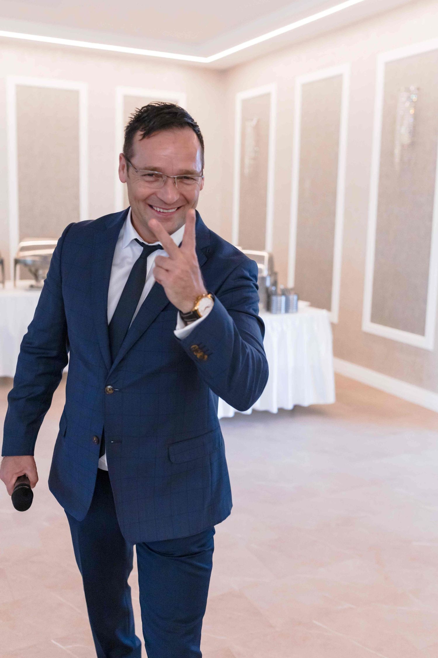A person in a navy blue suit laughs while leaning forward in a bright indoor event space.